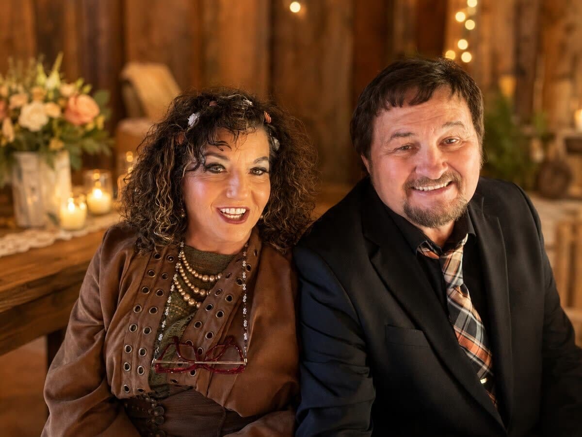 A middle-aged couple smiling together in a rustic, warmly lit room with a flower arrangement and candles in the background.