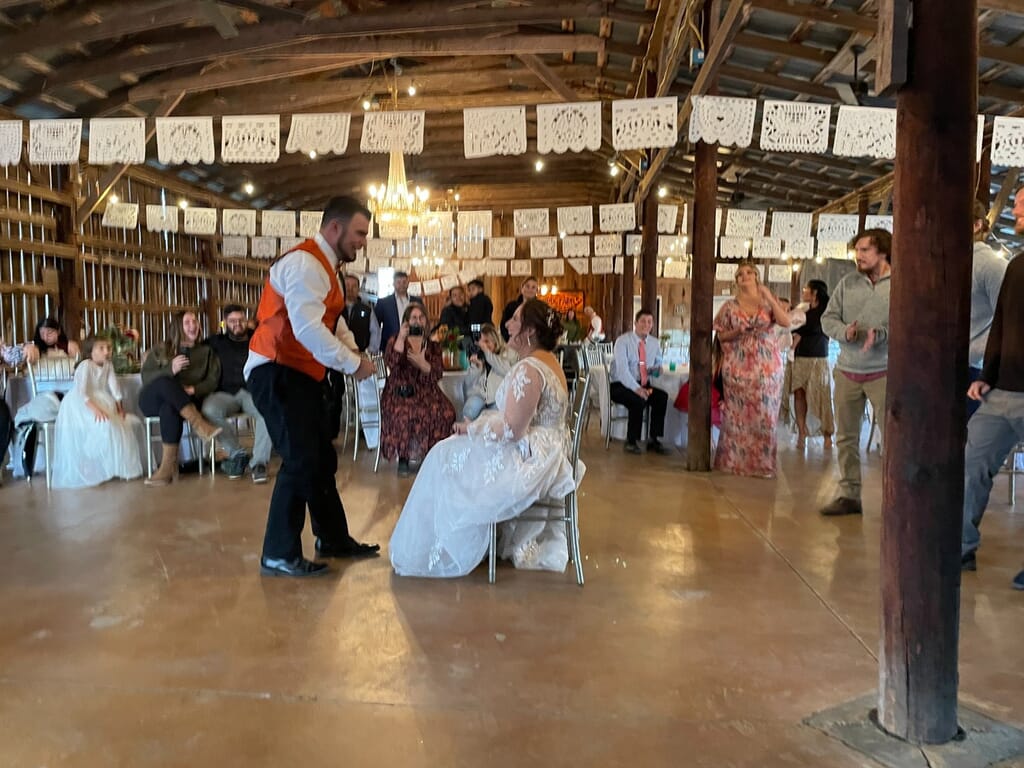 Burdoc Farms Wedding DJ | Crofton KY Wedding DJ & MC 13 A bride in a white gown sits in a chair as a groom in an orange vest stands before her at a rustic barn wedding reception