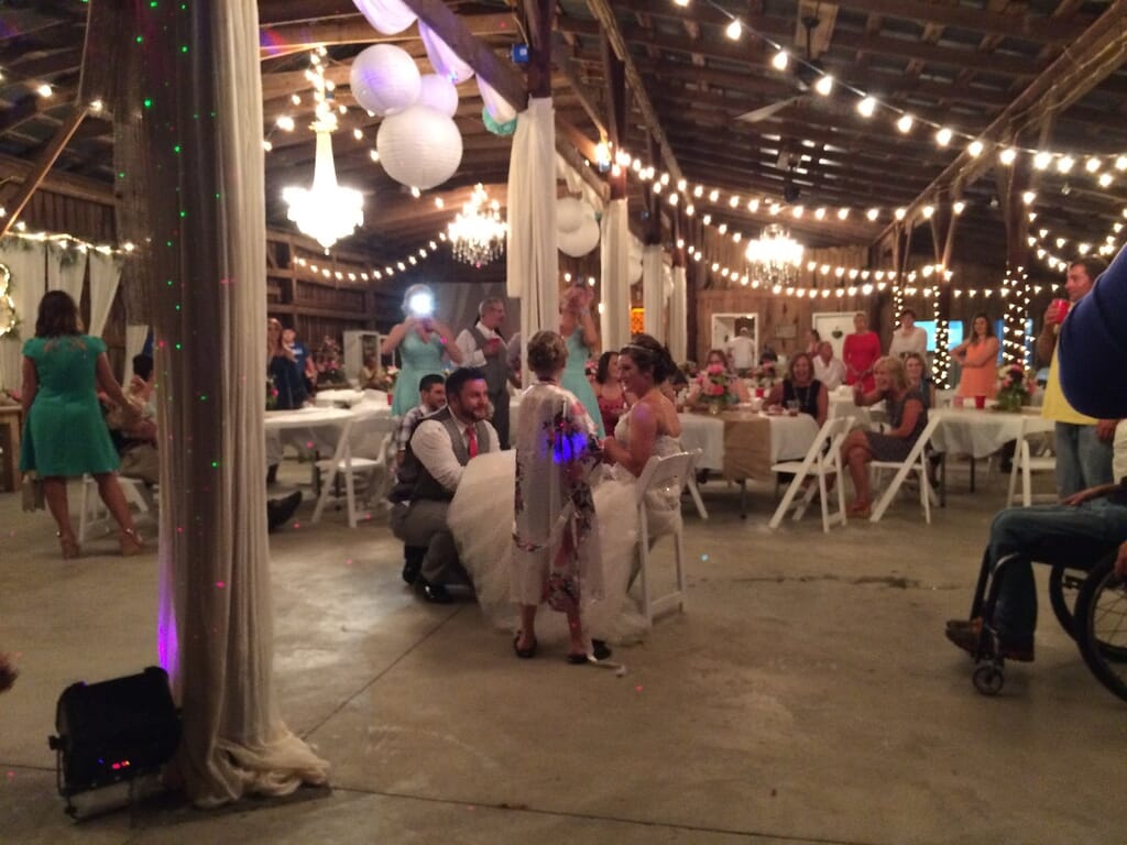 Burdoc Farms Wedding DJ | Crofton KY Wedding DJ & MC 10 A bride and groom sit in the center of a rustic barn reception, with guests gathered at tables decorated with lights.