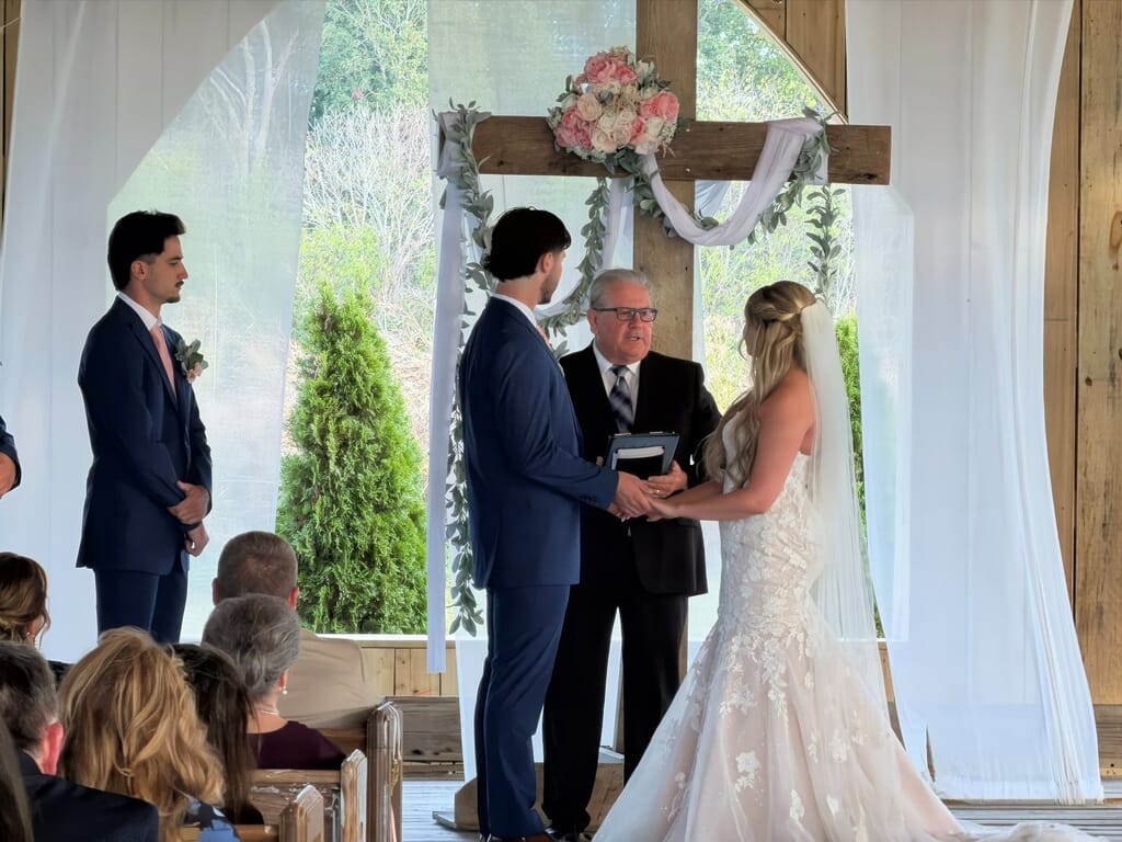 Burdoc Farms Wedding DJ | Crofton KY Wedding DJ & MC 4 A bride and groom hold hands during their wedding ceremony in front of a flower-decorated wooden cross