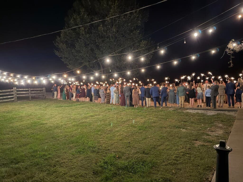 Burdoc Farms Wedding DJ | Crofton KY Wedding DJ & MC 3 Guests in formal attire hold lit sparklers in two lines on a lawn at night under string lights