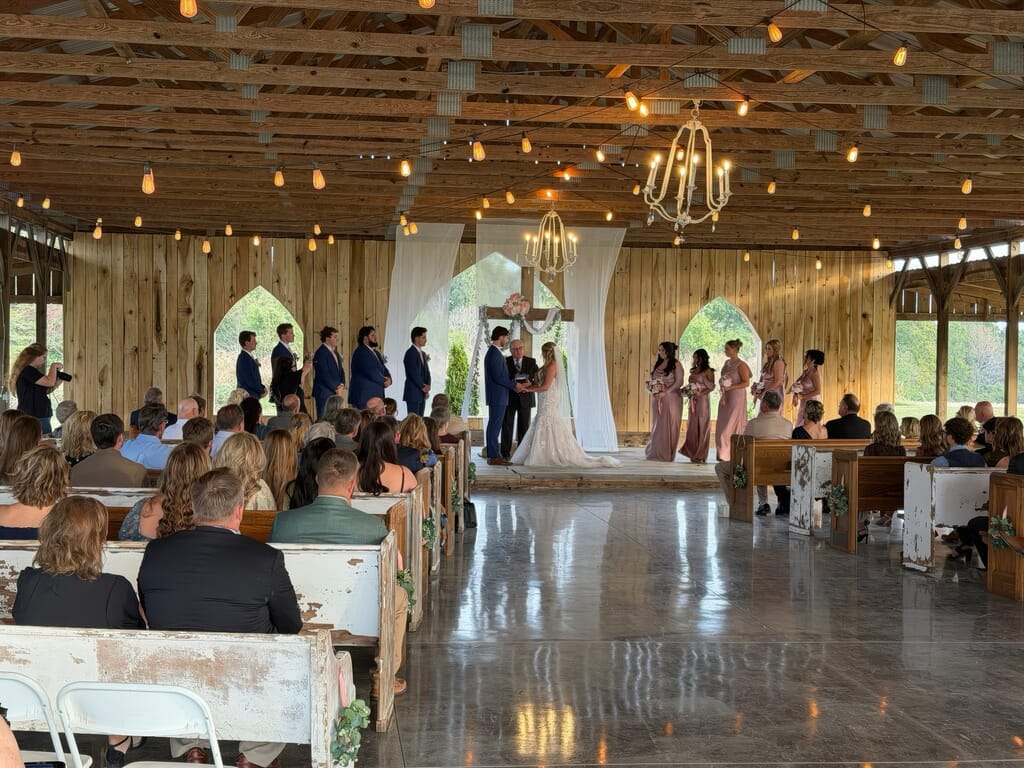 Burdoc Farms Wedding DJ | Crofton KY Wedding DJ & MC 1 A wedding ceremony in a rustic barn with a bride and groom at the altar, surrounded by their wedding party and guests