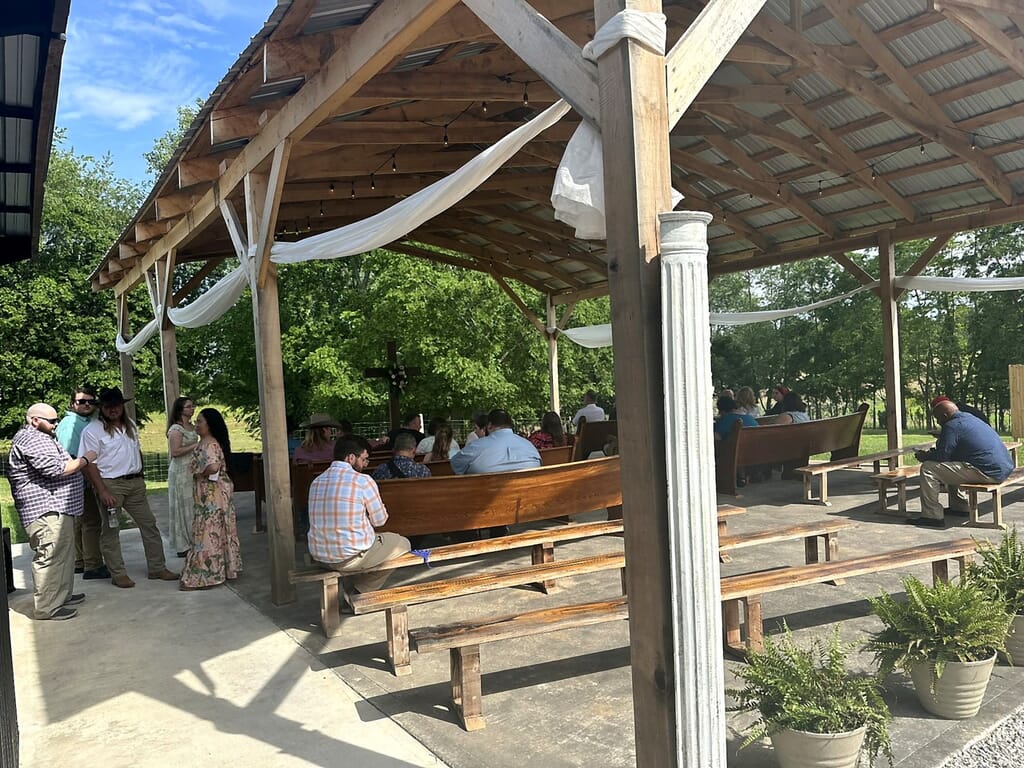 Wedding DJ Princeton Ky 9 Guests gather in an open-air wooden pavilion with pews and white fabric draping, with a wooden cross in the background