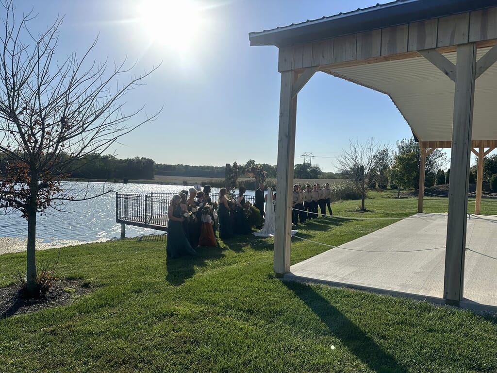 Newburgh Indiana Wedding DJ 3 A bride, groom, and wedding party stand before a floral arch for an outdoor wedding ceremony beside a lake