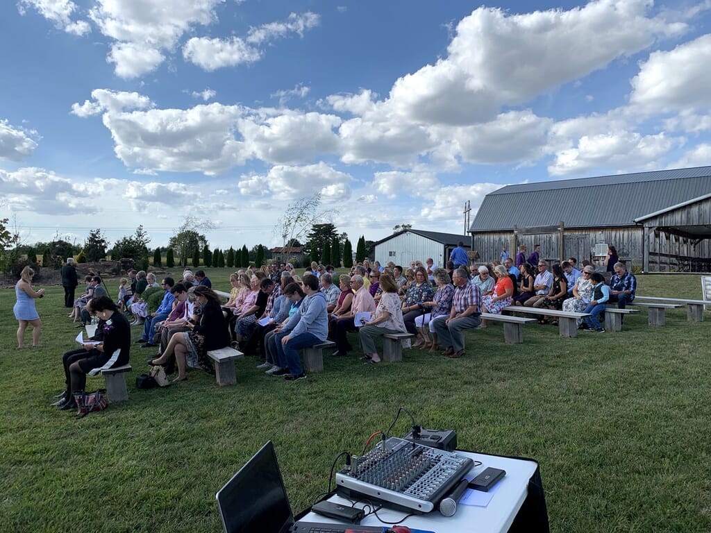 Newburgh Indiana Wedding DJ 5 Guests sit on wooden benches in a grassy field for an outdoor event, with a barn in the background and audio equipment in the foreground