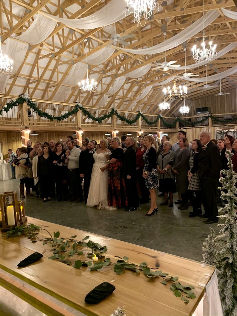 Newburgh Indiana Wedding DJ 4 A bride and groom stand with their wedding guests in a rustic barn decorated with white fabric and chandeliers