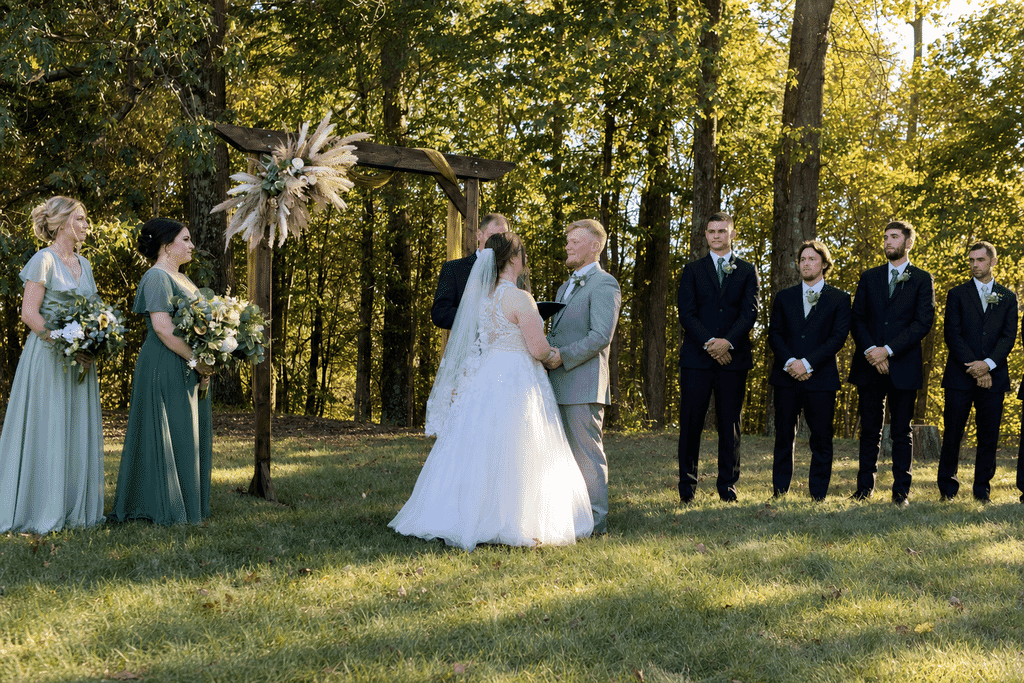 Central City Kentucky Wedding DJ 6 Bride and groom hold hands during an outdoor wedding ceremony under a wooden arch, flanked by bridesmaids and groomsmen