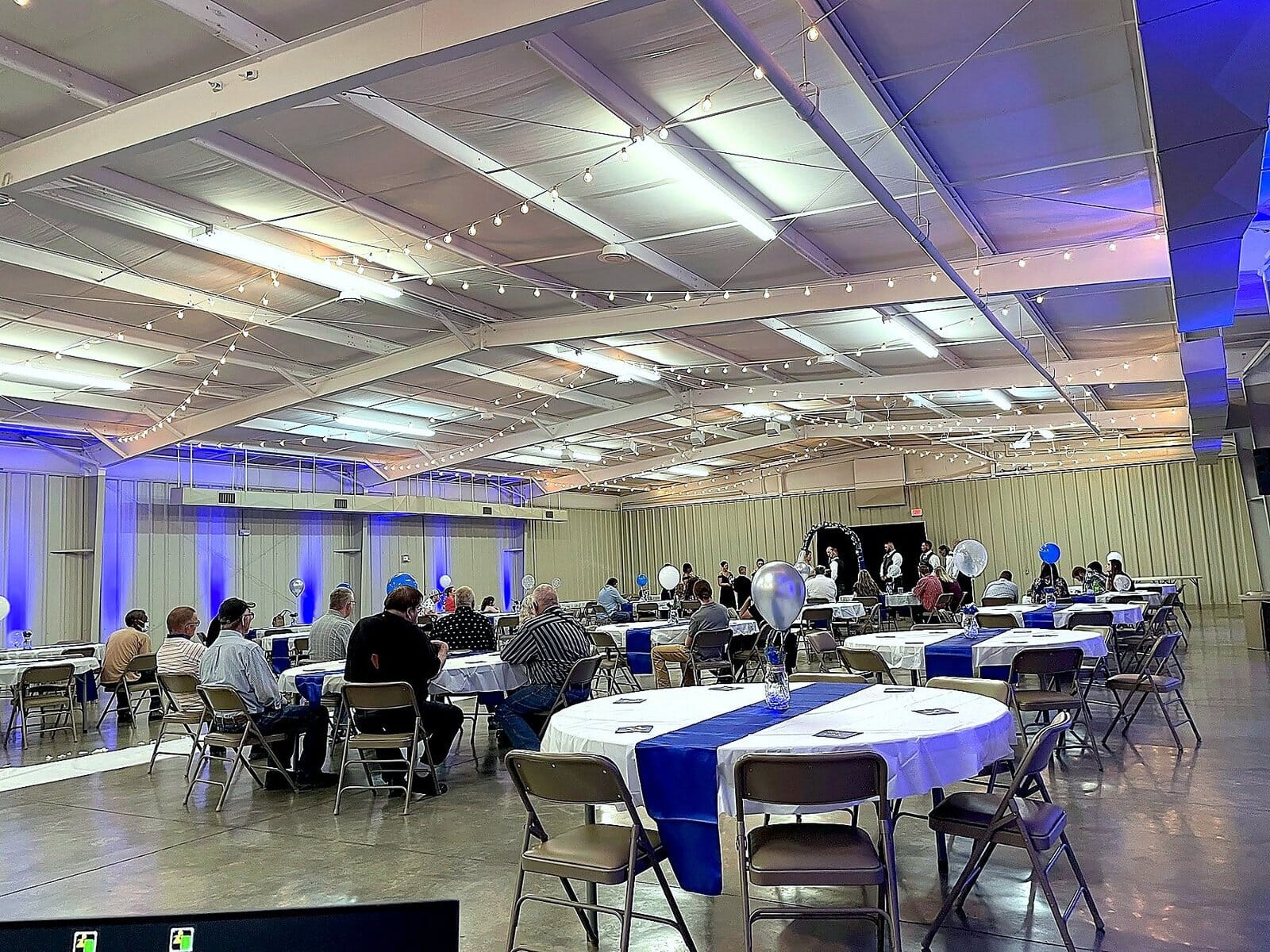 Event space with guests at round tables with blue and white linens under string lights and blue accent lighting.