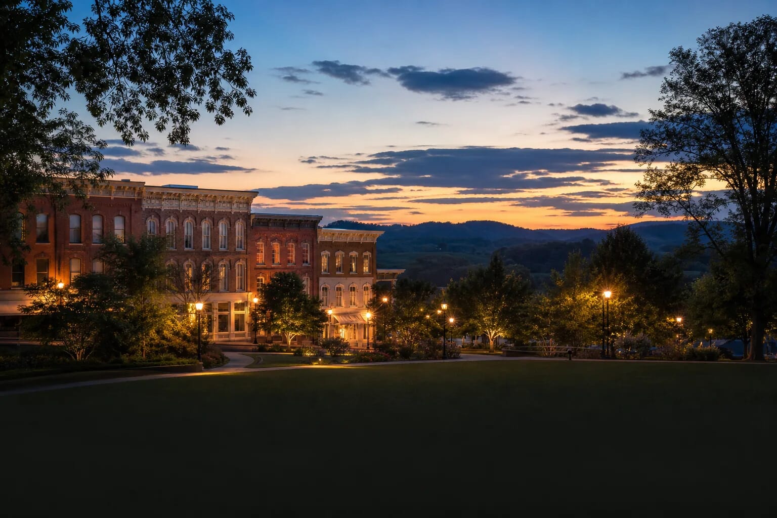 Historic brick buildings glow with warm window light along a park path during a colorful sunset over distant hills.