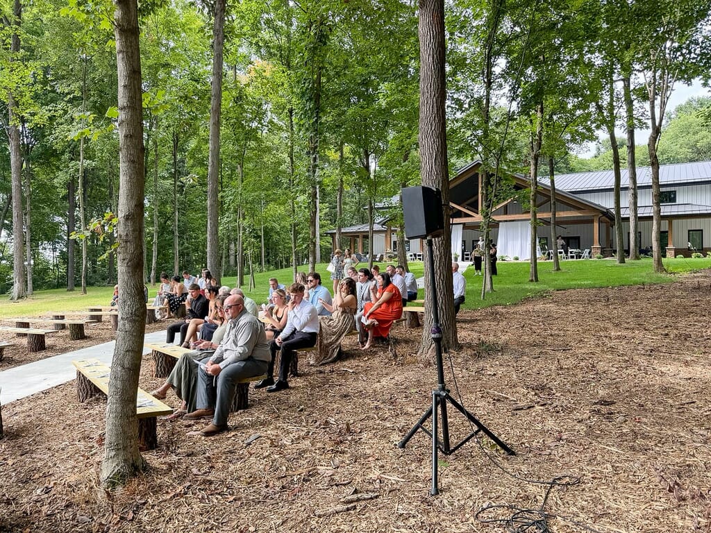 Gallery - Highbanks Wedding & Events Real Weddings 2 Guests seated on rustic wooden benches at the wooded outdoor ceremony site at Highbanks, with the venue building in the background.