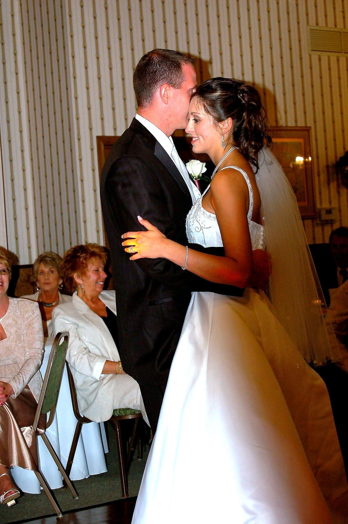 A groom in a tuxedo kisses the forehead of a bride in a white wedding dress as they dance together.