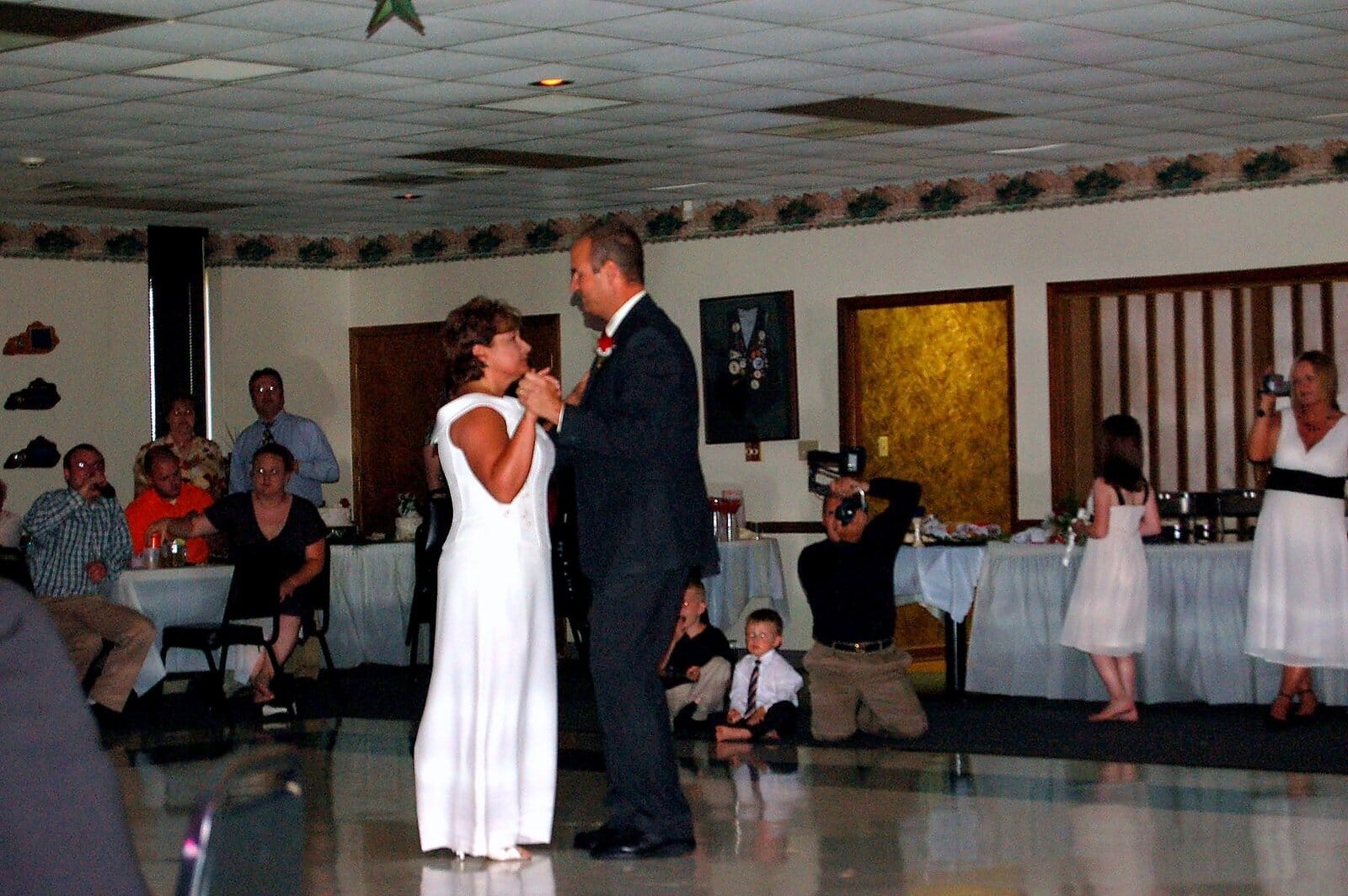 Bride and groom slow dancing at their wedding reception with guests and a videographer looking on.