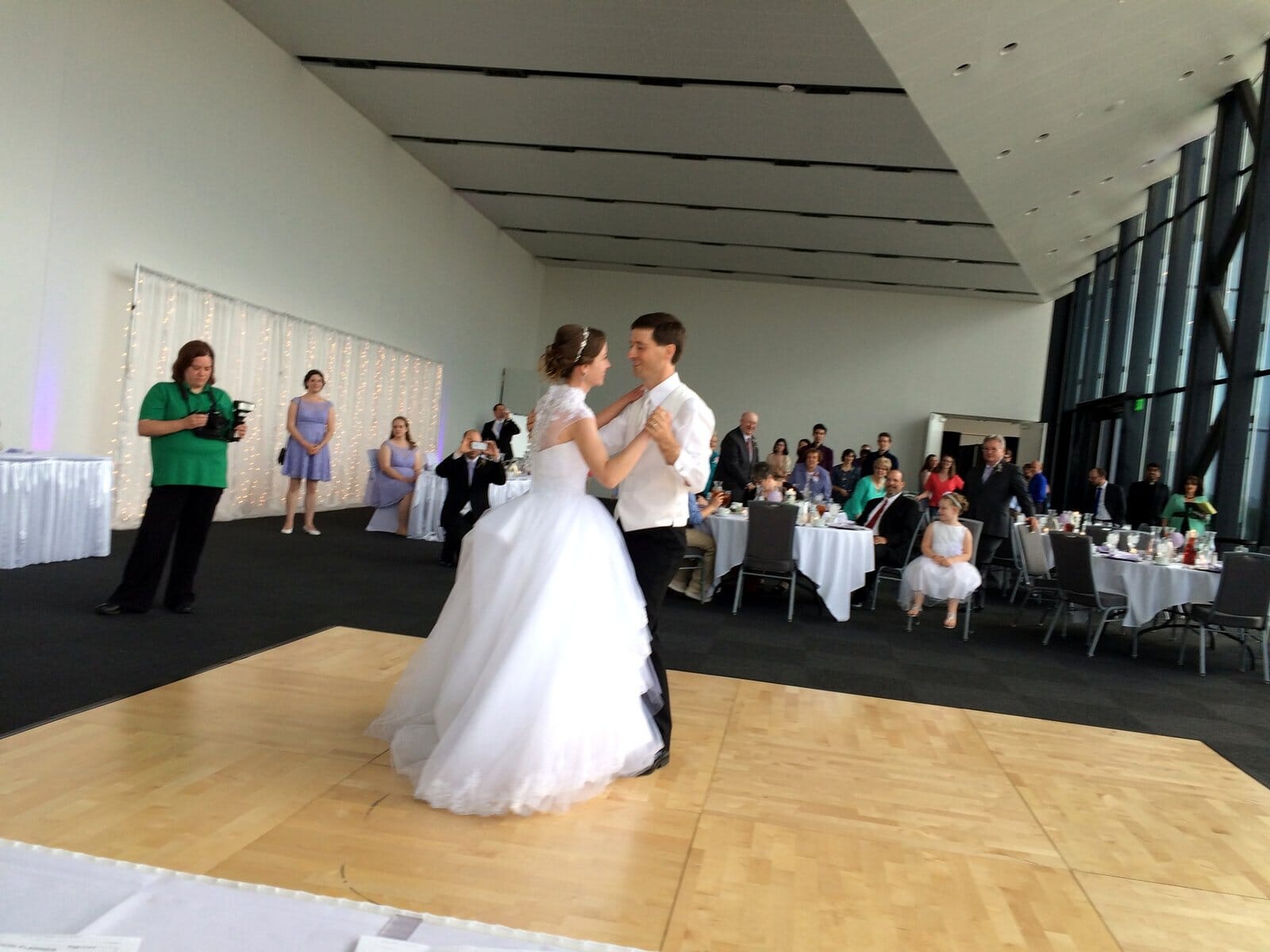 Bride and groom share their first dance on a wooden floor, surrounded by guests at a wedding reception.