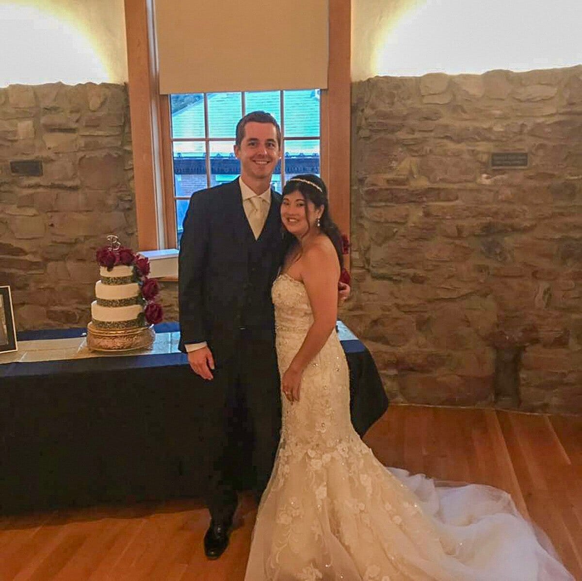 Smiling bride in a lace gown and groom in a dark suit standing by a wedding cake in a room with stone walls.