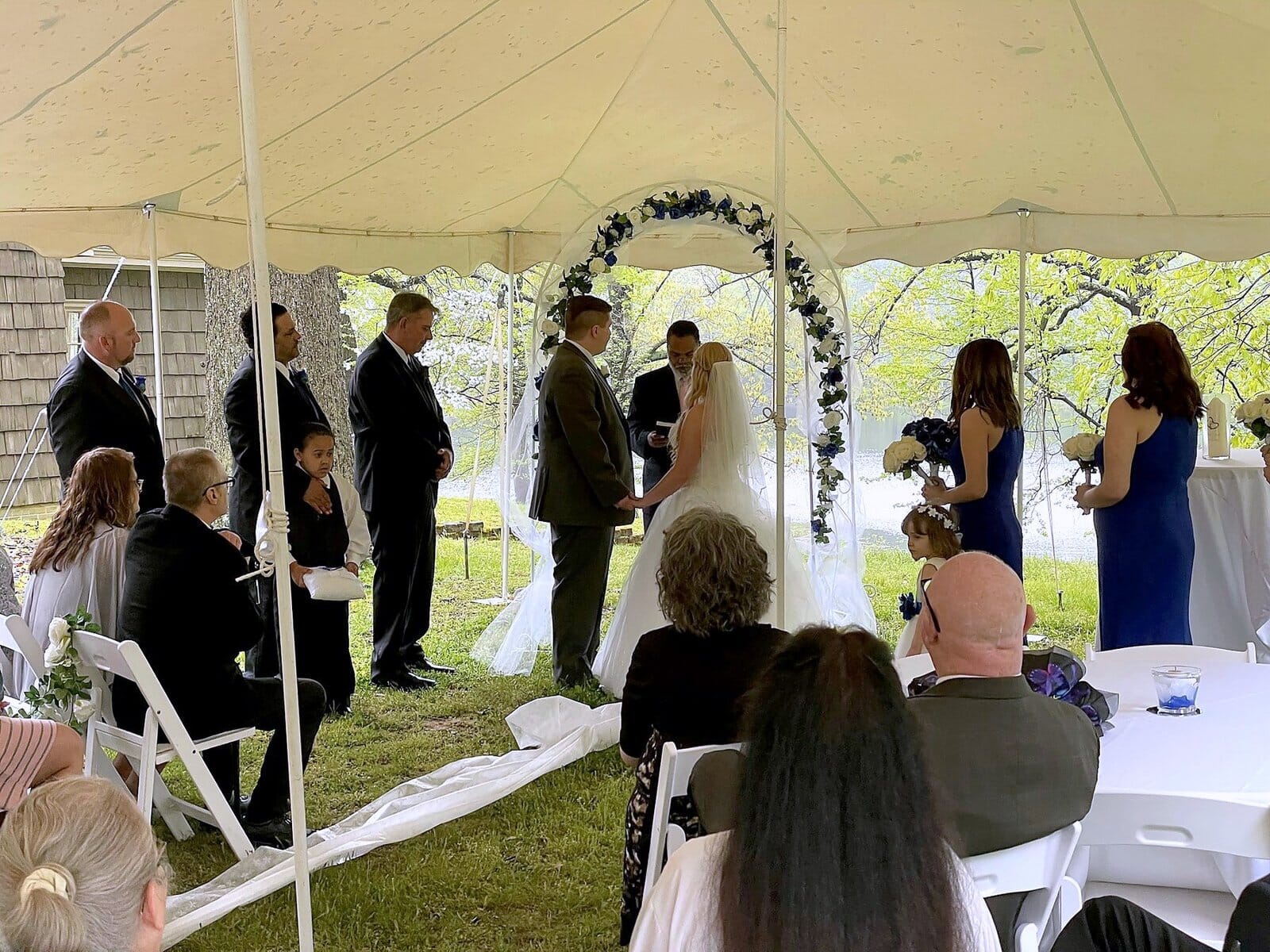 Wedding ceremony under a white tent with a bride and groom standing beneath a blue and white floral arch.